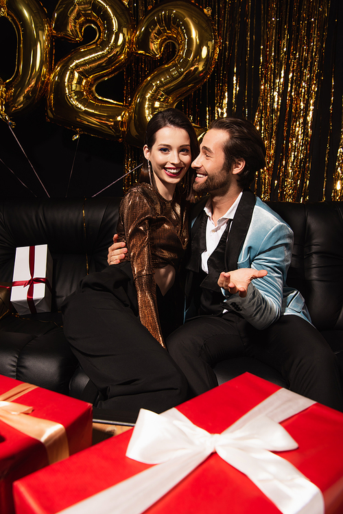 smiling man embracing elegant woman and pointing at blurred gift boxes near shiny christmas decoration on black