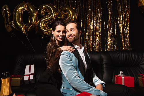 happy woman embracing elegant man on sofa near golden new year decoration on black