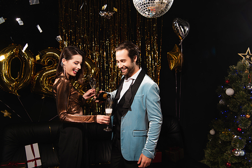 smiling man pouring champagne near elegant woman and shiny christmas decoration on black