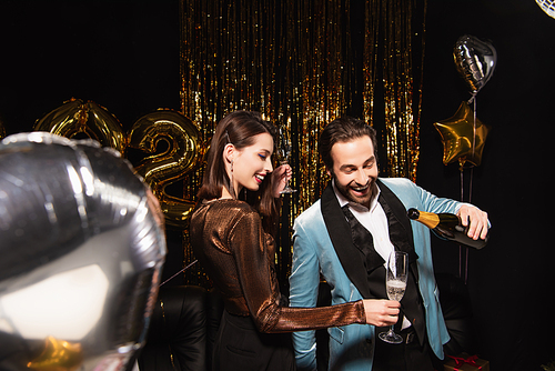 elegant woman holding glass near man pouring champagne while celebrating new year on black