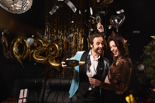 cheerful man pouring champagne near elegant woman during christmas party on black