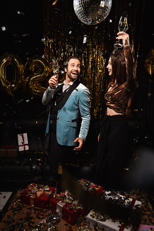 happy elegant couple toasting with champagne near golden christmas decoration on black