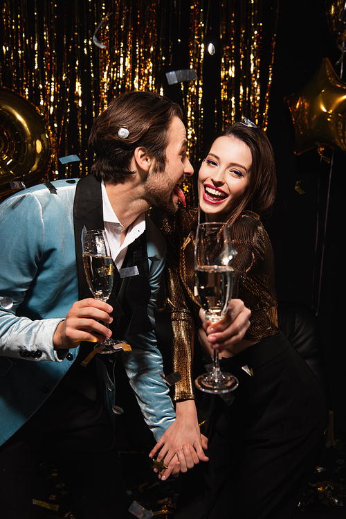 man sticking out tongue near cheerful woman during christmas party on black