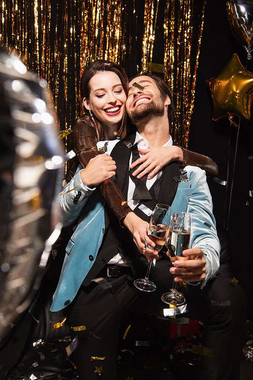 joyful and stylish couple clinking champagne glasses near sparkling christmas decoration on black