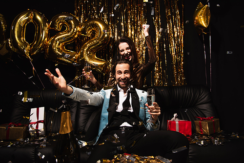 excited couple with champagne glasses showing yeah gesture near shiny decoration on black