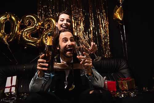 happy woman hugging man while clinking champagne glasses during christmas party on black