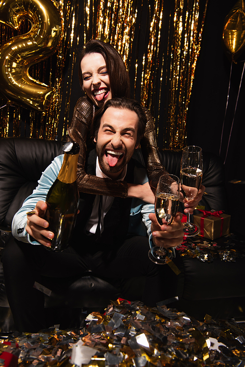 cheerful couple sticking out tongues while celebrating near year with champagne on black