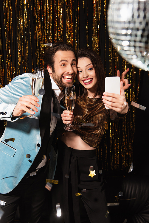 excited man showing victory gesture near woman taking selfie during new year party on black