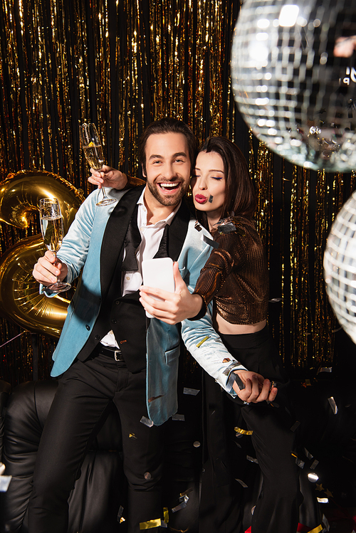 elegant woman pouting lips while taking selfie with cheerful man during christmas party on black