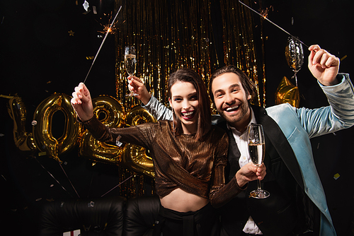 happy and elegant couple with sparklers and champagne  near golden decoration on black