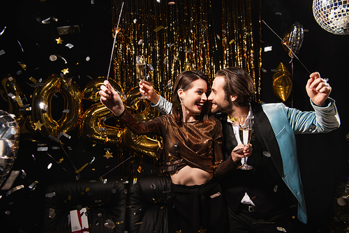 happy couple with sparklers and champagne smiling at each other during new year party on black