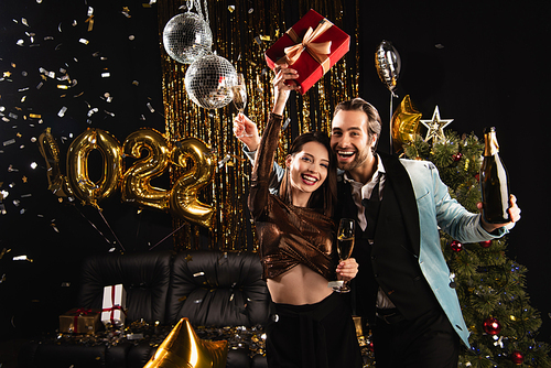 excited woman holding christmas present during festive party with elegant man on black