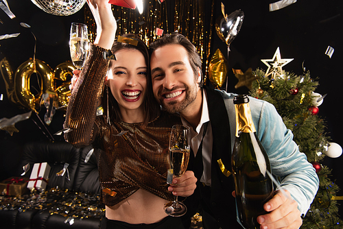 joyful couple with champagne smiling at camera near shiny christmas decoration on black