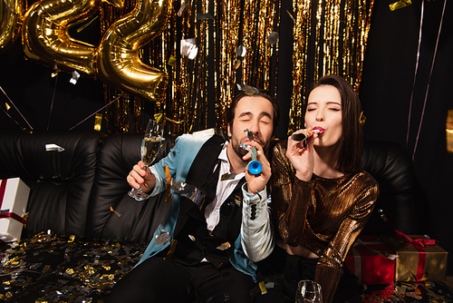 excited couple with champagne glasses blowing in party horns near sofa and christmas decoration on black