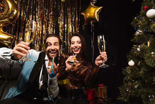 excited couple with champagne and party horns  near shiny christmas decoration on black