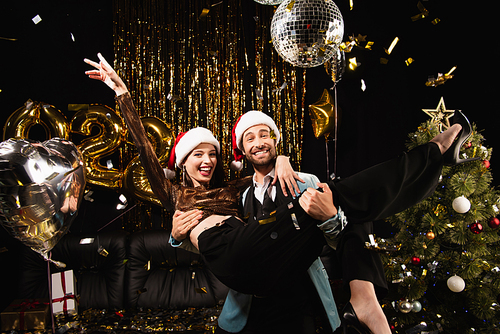 cheerful man in santa hat holding happy woman during sparkling christmas party on black