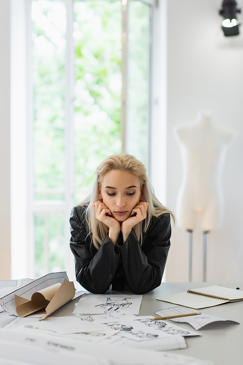 young fashion designer thinking near sewing patterns and sketches at workplace