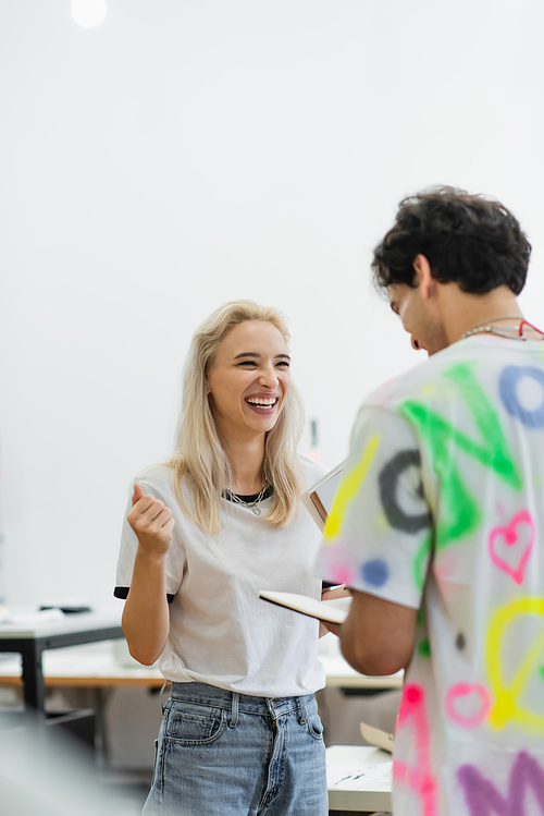 excited fashion designer laughing near colleague with notebook in tailor shop