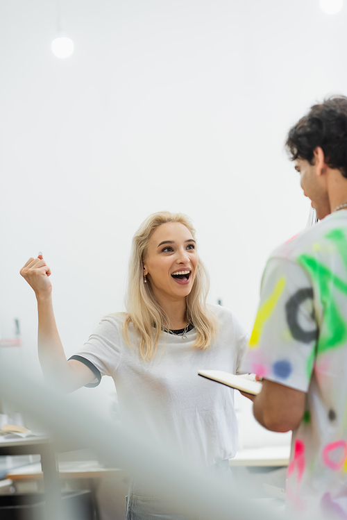 excited fashion designer showing win gesture near colleague, blurred foreground