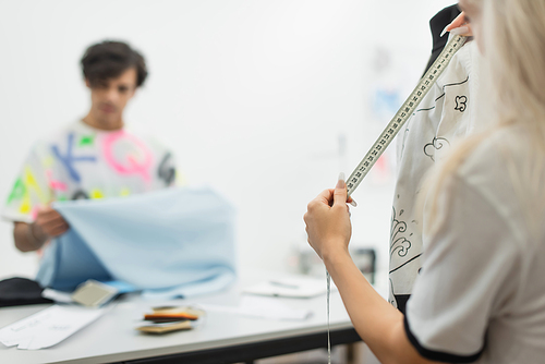 fashion designer measuring clothes on mannequin near blurred colleague holding tissue sample