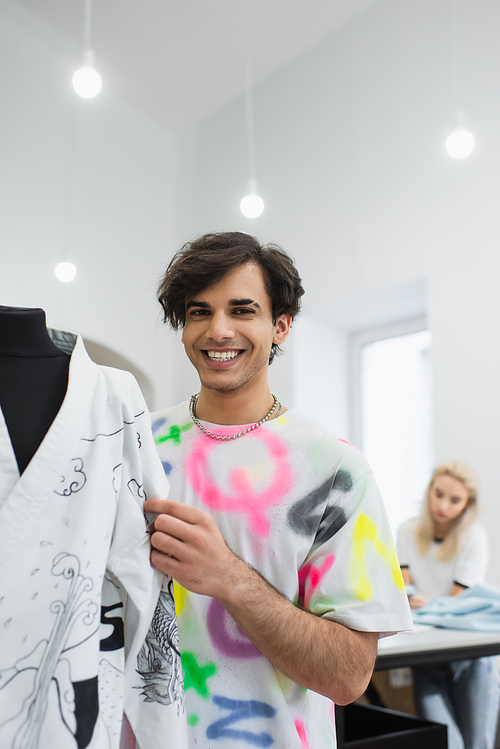 trendy designer smiling at camera near kimono on mannequin and colleague working on blurred background