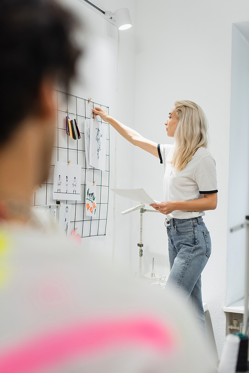 blonde designer attaching sketches on wall near blurred colleague