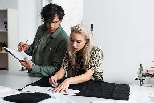 stylish designer writing in notebook near dressmaker cutting fabric in atelier