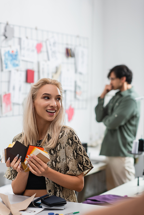 happy fashion designer holding color palette near colleague thinking near sketches on blurred background