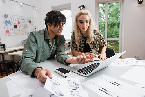 stylish designer pointing at laptop near blonde colleague and drawings at workplace