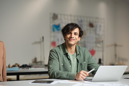 cheerful designer smiling at camera near laptop in fashion workshop