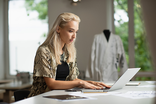blonde fashion designer typing on laptop near gadgets at workplace