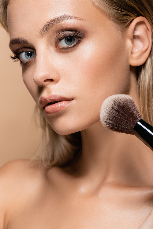 close up view of woman with perfect skin powdering face with soft cosmetic brush isolated on beige