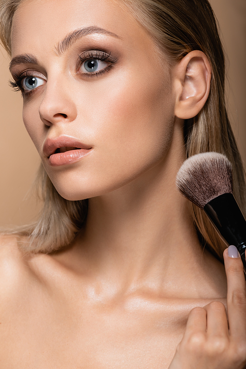 young woman with clean skin and grey eyes posing with large cosmetic brush isolated on beige