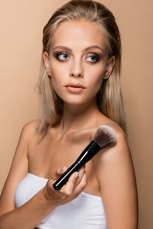 blonde woman with large and soft cosmetic brush looking away isolated on beige