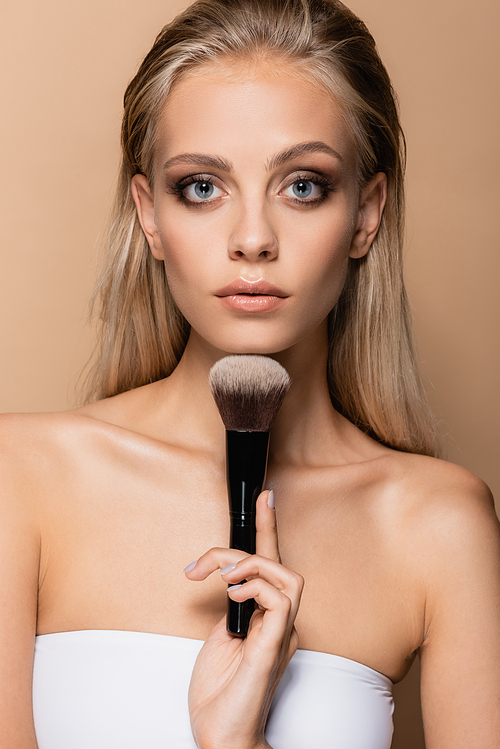 front view of woman with makeup holding large cosmetic brush near chin isolated on beige