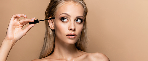 blonde woman with perfect skin and grey eyes holding black mascara isolated on beige, banner