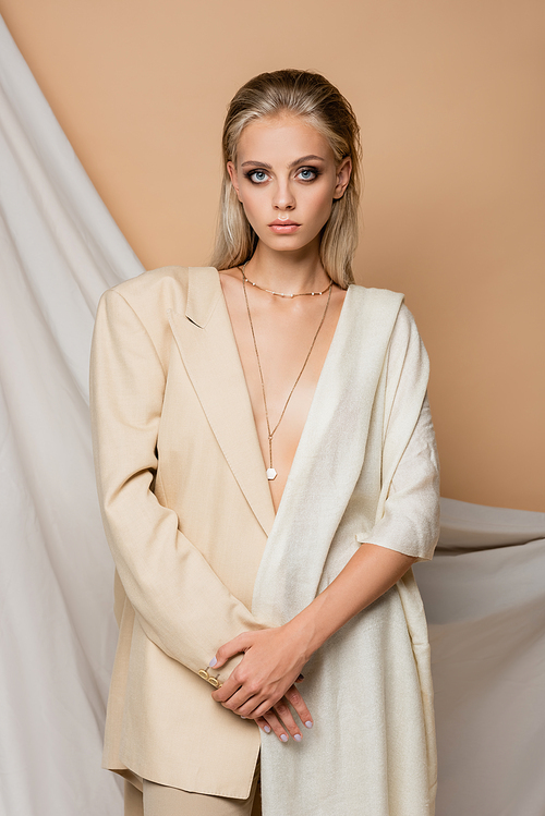 sensual woman in trendy blazer posing with shawl on shoulder near drapery on beige