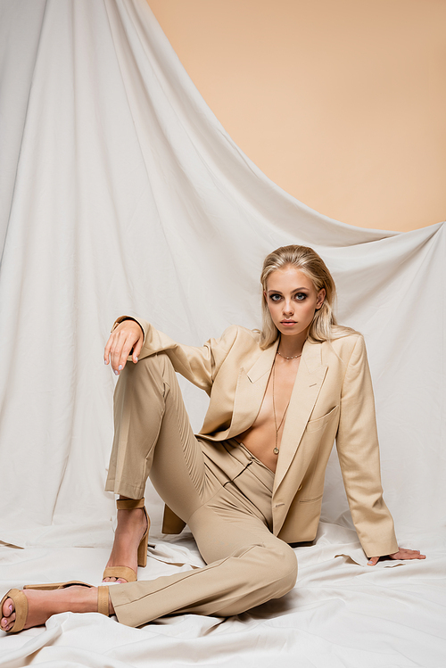 sensual woman in trendy suit  while sitting on beige background with drapery