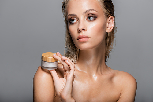 blonde woman with wet skin holding cosmetic cream near bare shoulder isolated on grey