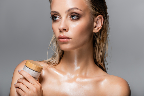 young woman with wet skin and hair holding glass jar of cosmetic cream isolated on grey