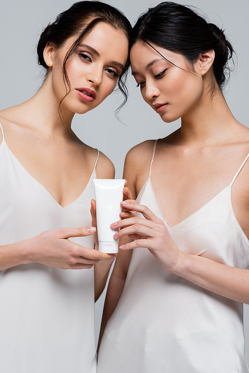 Young multiethnic women in silk dresses holding cosmetic cream isolated on grey