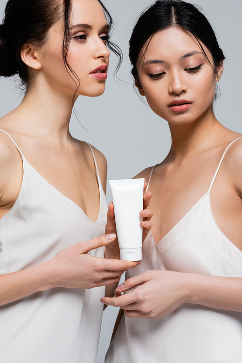 Pretty multiethnic women in silk dresses holding cream in tube isolated on grey