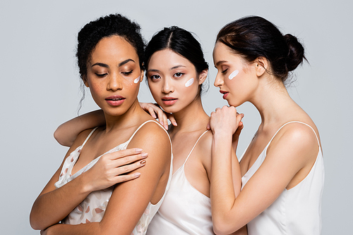 Multiethnic women with cosmetic cream on faces hugging each other isolated on grey