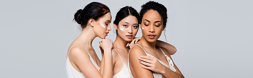 Young interracial women with cream on faces posing isolated on grey, banner