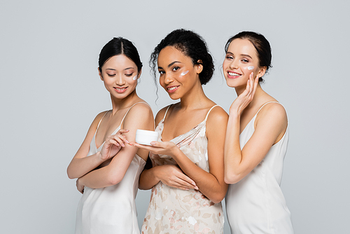 Positive african american woman holding container with cream near interracial friends isolated on grey