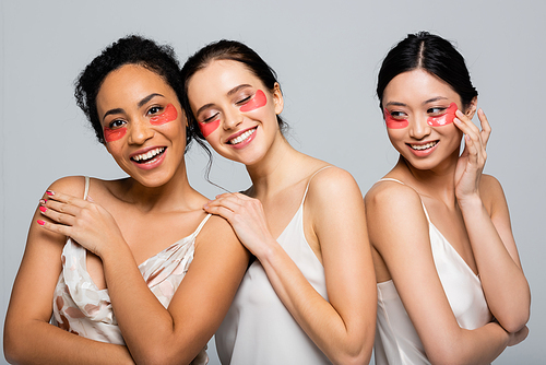Cheerful multicultural friends in eye patches and satin dresses posing isolated on grey