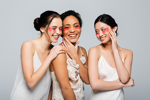 Cheerful multicultural women in silk dresses and eye patches isolated on grey