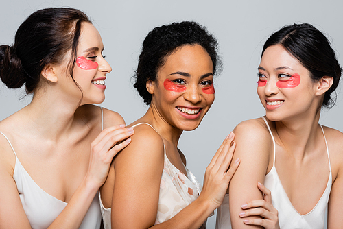 Happy interracial women with eye patches touching each other isolated on grey