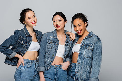 Smiling interracial women with red lips and denim jackets posing isolated on grey