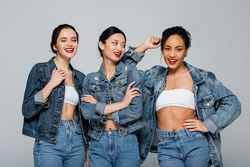 Happy interracial women with red lips and denim jackets posing isolated on grey